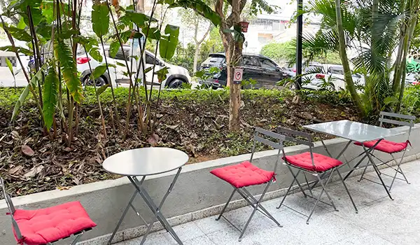 Imagem de uma área externa com mesas e cadeiras vermelhas ao lado de uma paisagem com árvores e carros estacionados na rua, ideal para momentos de descanso ao ar livre.