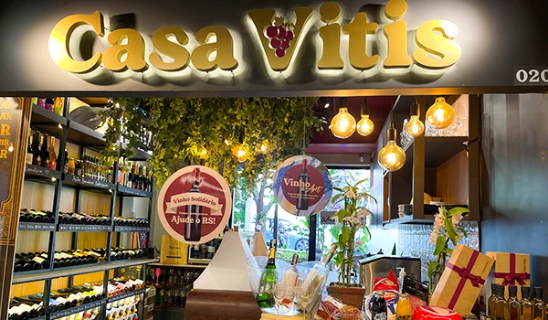Interior de uma loja de vinhos chamada Casa Vitis, com prateleiras cheias de garrafas de vinho, decoração moderna com lâmpadas pendentes e ambiente aconchegante.