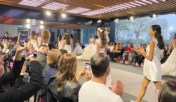 Desfile de moda com modelos usando vestidos de noiva, espectadores aplaudindo e registrando o evento com seus celulares em um ambiente elegante. Evento Casar.com para Noivas.