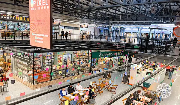 Imagem do Mercado de Santo Amaro com várias lojas abertas, pessoas sentadas em mesas e uma área com decoração colorida, promovendo comércio e lazer no centro comercial.