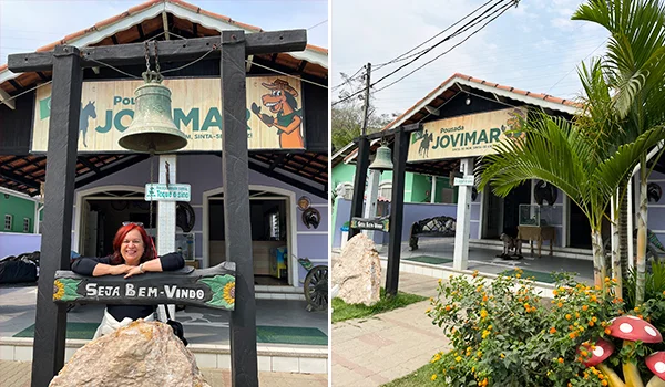 Fotografia da Pousada Jovimar, com fachada rústica, telhado de telhas cerâmicas e sino de bronze na entrada. À esquerda, uma mulher sorri sentada em um banco de madeira com a inscrição “Seja Bem-Vindo”. À direita, a entrada da pousada é cercada por plantas ornamentais, flores e um cogumelo decorativo.