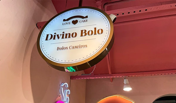 Vitrine de loja com painel arredondado exibindo o nome 'Divino Bolo' e a descrição 'Bolos Caseiros', destacando-se pela decoração em rosa e iluminação aconchegante.