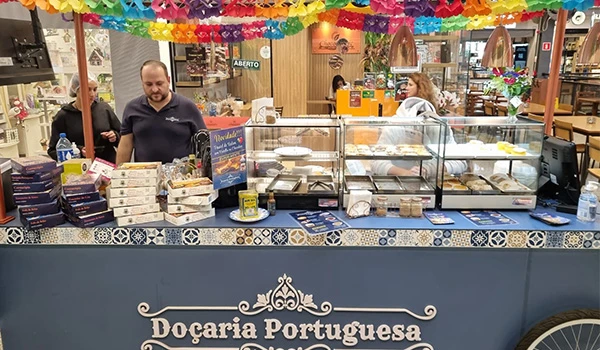 Imagem de uma banca de doces portugueses na Docaria Portuguesa, com diversas guloseimas e decoração colorida, atendida por funcionários sorridentes.
