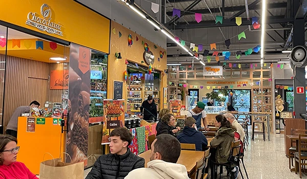 Interior do Empório 99 com clientes sentados em mesas, decorações coloridas, bandeirinhas e uma área de atendimento ao fundo com produtos variados. Mercado de Santo Amaro restaurantes.