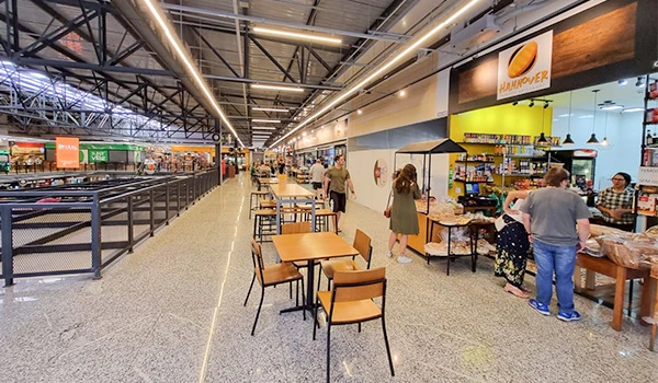 Imagem de uma praça de alimentação em um centro comercial, com várias mesas e cadeiras vazias, lojas ao fundo e pessoas fazendo compras ao redor.