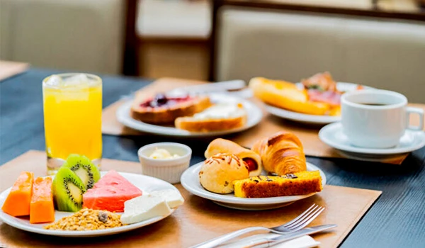 Delicioso café da manhã com frutas frescas, pães variados, e suco natural em uma mesa bem montada.