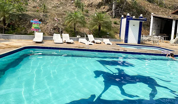 Piscina ao ar livre com água cristalina azul-turquesa, espreguiçadeiras brancas ao redor e uma área de lazer com ducha e mesas ao fundo, em meio a uma paisagem natural com vegetação e morros.