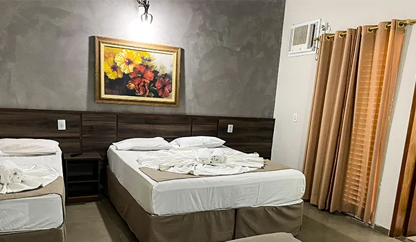 Quarto de pousada com duas camas de casal, roupas de cama brancas, cabeceira e mesas de apoio em madeira escura, quadro floral na parede cinza e cortinas marrons na janela.