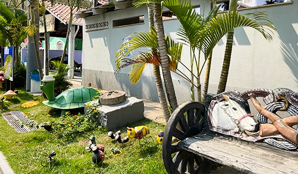 Área externa com jardim, palmeiras, plantas ornamentais e esculturas decorativas de tartaruga e animais, além de um banco rústico de madeira com rodas de carroça, em um ambiente ensolarado.