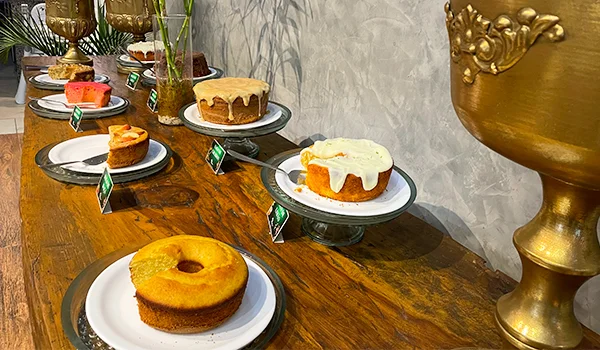 Mesa de café da manhã com diversos bolos caseiros dispostos em pratos sobre uma bancada de madeira, incluindo bolos simples, com calda de chocolate e cobertura branca, em um ambiente decorado com vasos dourados e plantas.