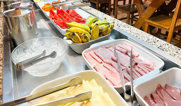 Mesa de café da manhã com bandejas de melancia, bananas, frios e queijos fatiados, além de gelo e recipientes de inox, em um ambiente com mesas e cadeiras de madeira.