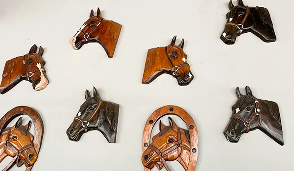 Decoração de parede com esculturas de cabeças de cavalos em madeira entalhada, em tons de marrom, preto e castanho, algumas com arreios e detalhes pintados, dispostas sobre uma parede branca.