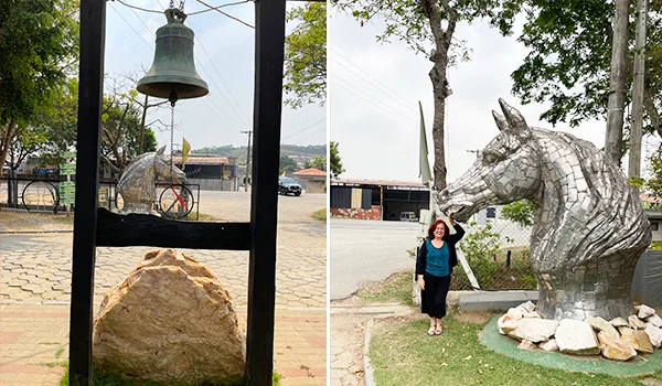 Decoração rústica em área externa, com um sino de metal suspenso em uma estrutura de madeira e uma escultura de cavalo feita com peças metálicas. Ao lado, uma visitante posa para a foto, em meio a árvores e elementos naturais.