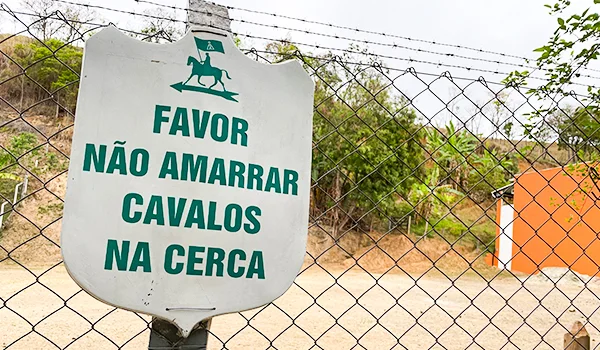 Placa branca com letras verdes em uma cerca de arame, com os dizeres “Favor não amarrar cavalos na cerca”. Ao fundo, há vegetação, chão de terra e uma construção laranja.