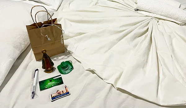 Cama com roupa de cama branca, sobre a qual estão dispostos uma pequena imagem religiosa, uma sacola de papel, um cartão de visita, uma caneta, um chaveiro e um cartão de acesso.