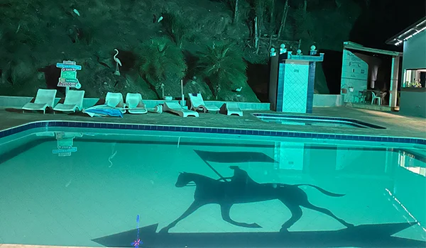 Piscina iluminada à noite, com a silhueta de um cavaleiro montado a cavalo pintada no fundo e refletida na água. Ao redor, há espreguiçadeiras, vegetação tropical e uma área de lazer.