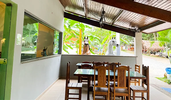 Restaurante rústico e arejado, com mesas e cadeiras de madeira, localizado em uma varanda coberta com vista para a área verde e cavalos ao fundo.