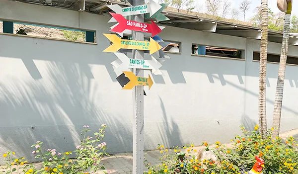 Placas coloridas de sinalização turística indicando cidades como São Paulo, Santos e Campos do Jordão, instaladas em um jardim com flores e palmeiras diante de um muro cinza.