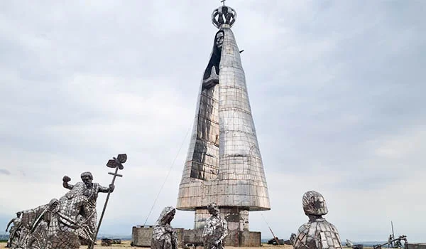 Monumento metálico em forma de cone, com uma coroa no topo e esculturas de figuras humanas ao redor, instalado em uma área aberta sob um céu nublado.