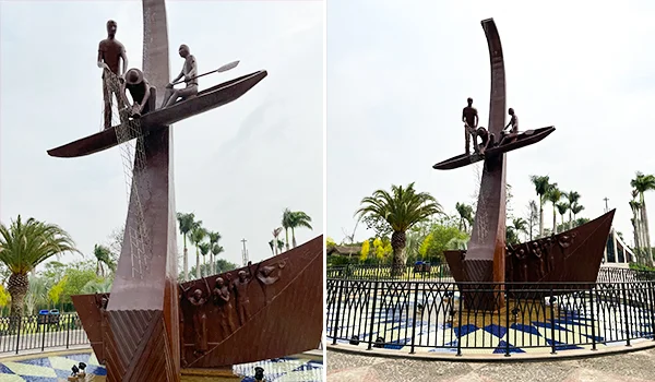 Monumento aos Três Pescadores, com esculturas de pescadores em uma canoa e redes de pesca sobre uma estrutura vertical, instalado em uma praça ajardinada, cercado por grades, palmeiras e vegetação.