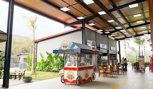 Área de alimentação do Parque Três Pescadores, com carrinho de pipoca gourmet, quiosques, mesas e cadeiras de madeira sob uma ampla cobertura, cercada por jardins e plantas tropicais.