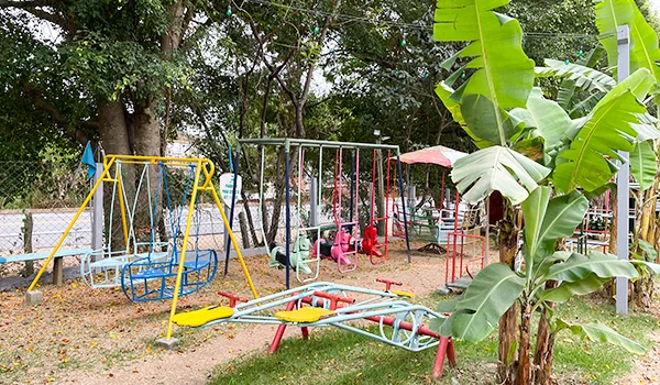 Parquinho ao ar livre com balanços, gangorras e brinquedos de metal, cercado por árvores, vegetação e bananeiras, em uma área gramada com folhas espalhadas pelo chão.