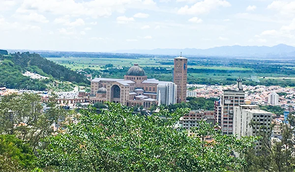Vista panorâmica do Santuário Nacional de Aparecida, com sua cúpula e torre de destaque, cercado pela área urbana, vegetação e montanhas ao fundo, sob um céu parcialmente nublado.