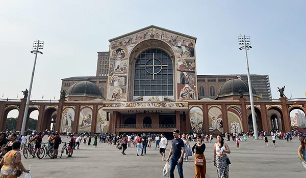 Fachada do Santuário Nacional de Aparecida, com grandes painéis de mosaicos religiosos, vitrais e arcos, enquanto visitantes circulam pela ampla praça em frente à basílica.