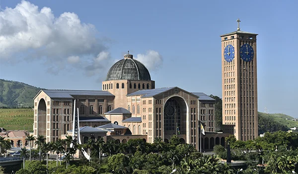 Vista externa do Santuário Nacional de Aparecida, com sua arquitetura monumental, cúpula central, torre alta com relógios azuis e cruz no topo, cercado por jardins e vegetação, sob um céu azul com nuvens.