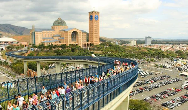 Vista panorâmica do Santuário Nacional de Aparecida, com sua cúpula e torre dos relógios ao fundo, enquanto uma passarela curva elevada reúne peregrinos e visitantes. Ao lado, há um amplo estacionamento com diversos veículos e a paisagem urbana da cidade.