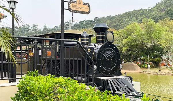Locomotiva a vapor preta em exposição junto a um lago, cercada por vegetação exuberante e montanhas arborizadas, em um ambiente turístico ao ar livre.