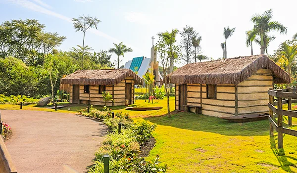 Réplicas de casas tradicionais de pescadores, com paredes de barro e madeira e telhados de palha, em uma área ajardinada com caminho pavimentado, gramado, flores e palmeiras.