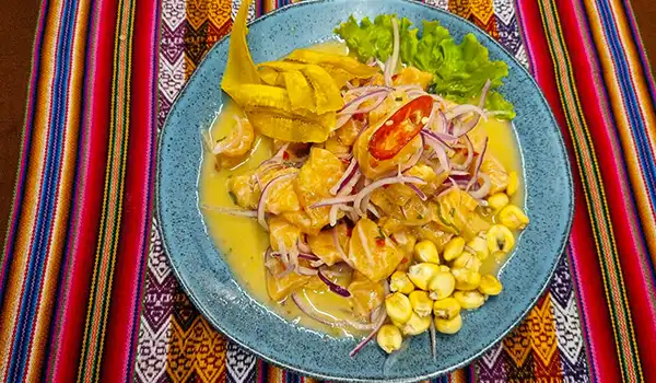Prato de ceviche peruano com peixe branco, cebola, pimentão, milho, e limão, servido em um prato azul decorado com folhas de alface, sobre pano colorido tradicional.