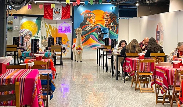 Ambiente de restaurante com decoração colorida e mesas com toalhas de mesa rosa, com pessoas jantando e uma parede com pintura de dois personagens indígenas.