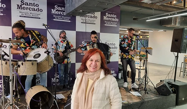 Conjunto musical ao vivo no Mercado de Santo Amaro, com músicos tocando instrumentos e Deusa Rodrigues na frente do palco, promovendo entretenimento local.