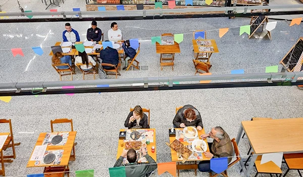 Cena de uma Mercado de Santo Amaro com várias bancas, pessoas fazendo compras, organizadores e clientes interagindo, decorada com bandeirinhas coloridas, vista ampla do ambiente.