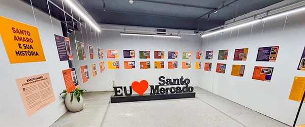 Exposição sobre a história do santo amor e cultura do mercado em sala de museu com painéis coloridos e símbolo 'EU❤Santo Mercado' no centro.
