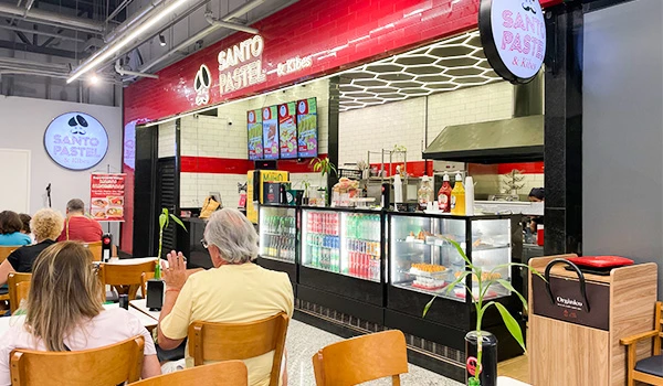 Interior de uma lanchonete na o Santo Pastel com clientes sentados enquanto fazem pedidos em caixa, decorada com cartazes coloridos e iluminação moderna. Mercado de Santo Amaro restaurantes.