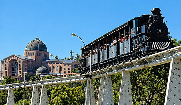 Locomotiva a vapor preta com passageiros atravessando uma ponte metálica elevada, diante de uma igreja com cúpula, sob um céu azul e com vegetação ao fundo.