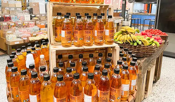 Vários frascos de mel de produção artesanal em embalagem de vidro na loja, ao lado de uma cesta com bananas e outras frutas, destaque para o mel dourado e embalagem informativa.