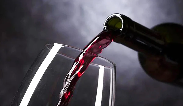 Vinho sendo servido em uma taça transparente, com destaque para a bebida de cor vermelha, e uma garrafa inclinada no topo da taça.