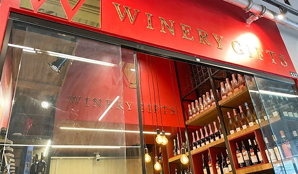 Fachada de uma loja de vinhos com baldes de vinho em prateleiras na vitrine, decorada com iluminação moderna e marca 'Winery Gifts'