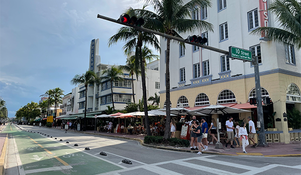 Uma vista vibrante da 10th Street em Miami Beach, com palmeiras, tráfego e restaurantes ao ar livre, ideal para mostrar o estilo de vida da Flórida.