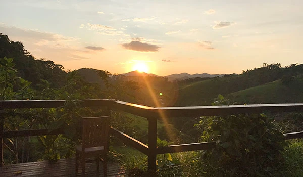 Vista de um pôr do sol em um mirante com cadeiras de madeira e uma paisagem de montanhas verdes ao fundo, ideal para relaxar e apreciar a natureza.