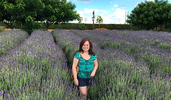 Deusa Rodrigues no campo de lavanda ao ar livre, com árvores ao fundo e céu azul, em um dia ensolarado, vestindo camiseta verde e shorts pretos. Lugares floridos em São Paulo.