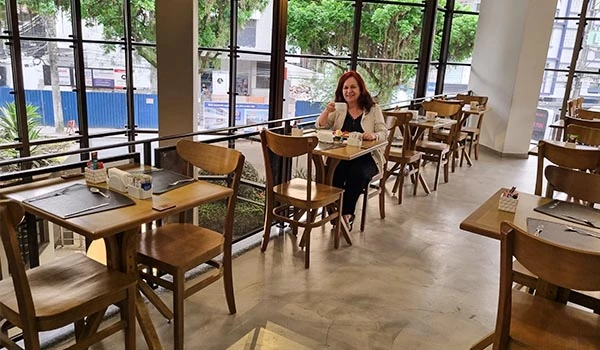Deusa Rodrigues numa mesa de restaurante com várias mesas e cadeiras de madeira, ambiente iluminado por grandes janelas com visão de árvores e rua.