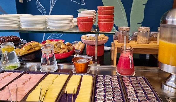 Buffet de café da manhã com várias opções de alimentos doces e salgados, incluindo geleias, queijos, frutas, pães e itens de café. Ambiente colorido e bem organizado.