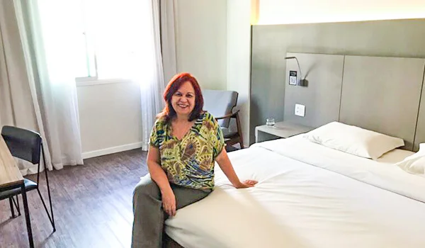 Foto de Deusa Rodrigues na cama de hotel na região do Ibirapuera. Onde se nota cama com lençóis brancos e tons de verde nas paredes.