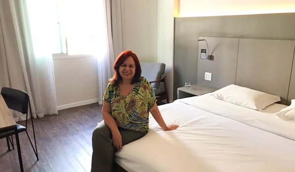Deusa Rodrigues sentada na beirada de uma cama de hotel, com iluminação natural vindo de uma janela ao lado, decoração moderna, evidenciando conforto e hospitalidade.