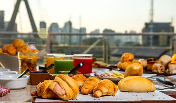 Foto do café da manhã no Novotel São Paulo Morumbi, uma mesa com pães, frios, sucos, doces e ao fundo vidraça onde se observra a Ponte Estraiada.